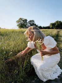 Robe en dentelle blanche avec des manches bouffantes, jupe froncée ; femme cueillant des fleurs sauvages dans un champ herbeux sous un ciel bleu clair.