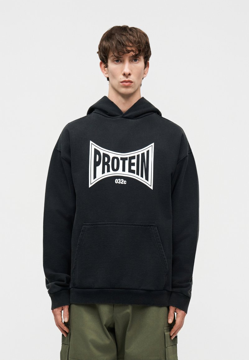 Jonge man die een zwarte hoodie draagt met witte "PROTEIN 032c"-logo en olijfgroene cargobroek, staand tegen een effen witte achtergrond.