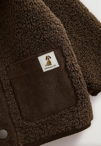 Brun strukturerad fleece med ett manchestertygsficka med en hundpatch och texten "AUTHENTIC ME" på ett klädesplagg.