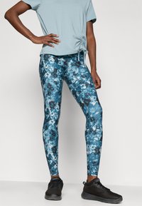 Zilās ziedu leggingas ar gludu tekstūru un elastīgu piegādi, saskaņotas ar gaiši zilu blūzi ar sānu rušinājumu un melniem sporta apaviem.