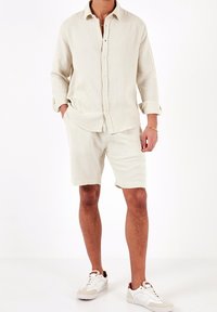 Camicia beige con bottoni e maniche arrotolate, abbinata a pantaloni corti beige coordinati, entrambi in tessuto testurizzato. Indossata con sneakers bianche.