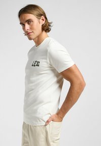 Camiseta de algodón blanca con un logo en bloque impreso en verde "LEE" en el lado izquierdo del pecho. Mangas cortas y escote redondo.
