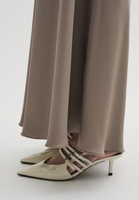 Beige patentlæderhæle med spidse tæer og tynde stilethæle. Remme over vristen og blødt beige stof, der hænger ned fra nedenunder.