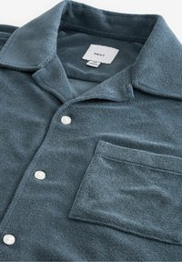 Tummansininen, pehmeäpintainen napitettava paita, jossa kaulus, rintatasku ja kaksi valkoista nappia edessä. Bränditunniste näkyvissä.