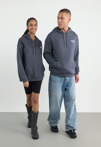 Gray hoodies with front pockets, featuring logo detail. Woman wears black shorts and knee-high black boots; man in faded baggy jeans and black shoes.