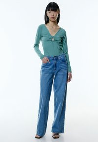 Mintgroene geribbelde lange mouw top met een centrale sleutelgat, gecombineerd met hoge lichtblauwe wijde jeans en zwarte haksandalen.