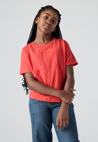 Camiseta de algodón roja con el logo "LEVI'S" emblema, cuello redondo, mangas cortas y un corte relajado. Combinada con jeans de mezclilla azul.