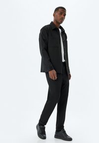 Camisa negra de botones con dos bolsillos en el pecho, combinada con pantalones negros ajustados y zapatillas negras, sobre un fondo blanco liso.