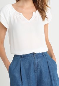 Kvinna som bär en vit kortärmad blus med volangkrage, instoppad i höga blåjeans, med händerna i fickorna.