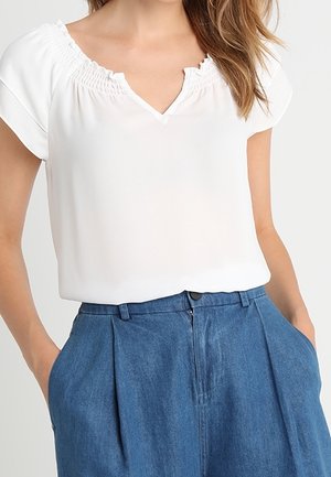 Donna con una blusa bianca a maniche corte e scollatura volant infilata in un paio di pantaloni in denim blu a vita alta, con le mani in tasca.