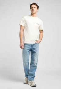 Ljusbeige bomullströja med en liten LEE-logotyp på bröstet, tillsammans med ljusblå denimjeans och grå sportskor.