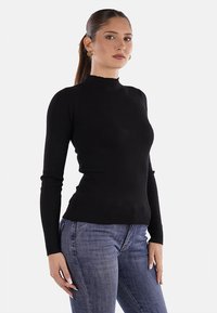 Top lungo a maniche lunghe ribbed nero con collo alto, silhouette aderente e tessuto testurizzato, abbinato a jeans blu.