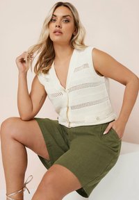 Stickad krämfärgad väst med hålstickad design, ihop med olivgröna shorts. Vesten har en djup V-ringning och knapppdetaljer.
