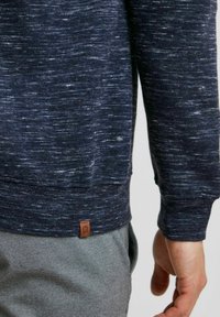 Pull bleu marine avec un tricot texturé, présentant un ourlet et des poignets côtelés. Une petite étiquette en cuir de marque est visible à l'ourlet inférieur.