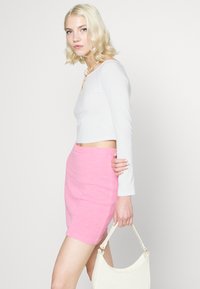 Top cropped branco de manga longa combinado com uma saia mini texturizada rosa. O modelo segura uma carteira branca, exibindo um design simples.