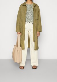 Oliwkowa długa kurtka noszona na wzorzystej zielono-białej bluzce, w zestawieniu z białymi spodniami o szerokich nogawkach, brązowymi sandałami i beżową torebką.