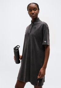 Žena v voľných tmavosivých šatách typu polo, drží čiernu fľašu na vodu a pozerá sa do boku na jednoduchom svetlom pozadí.