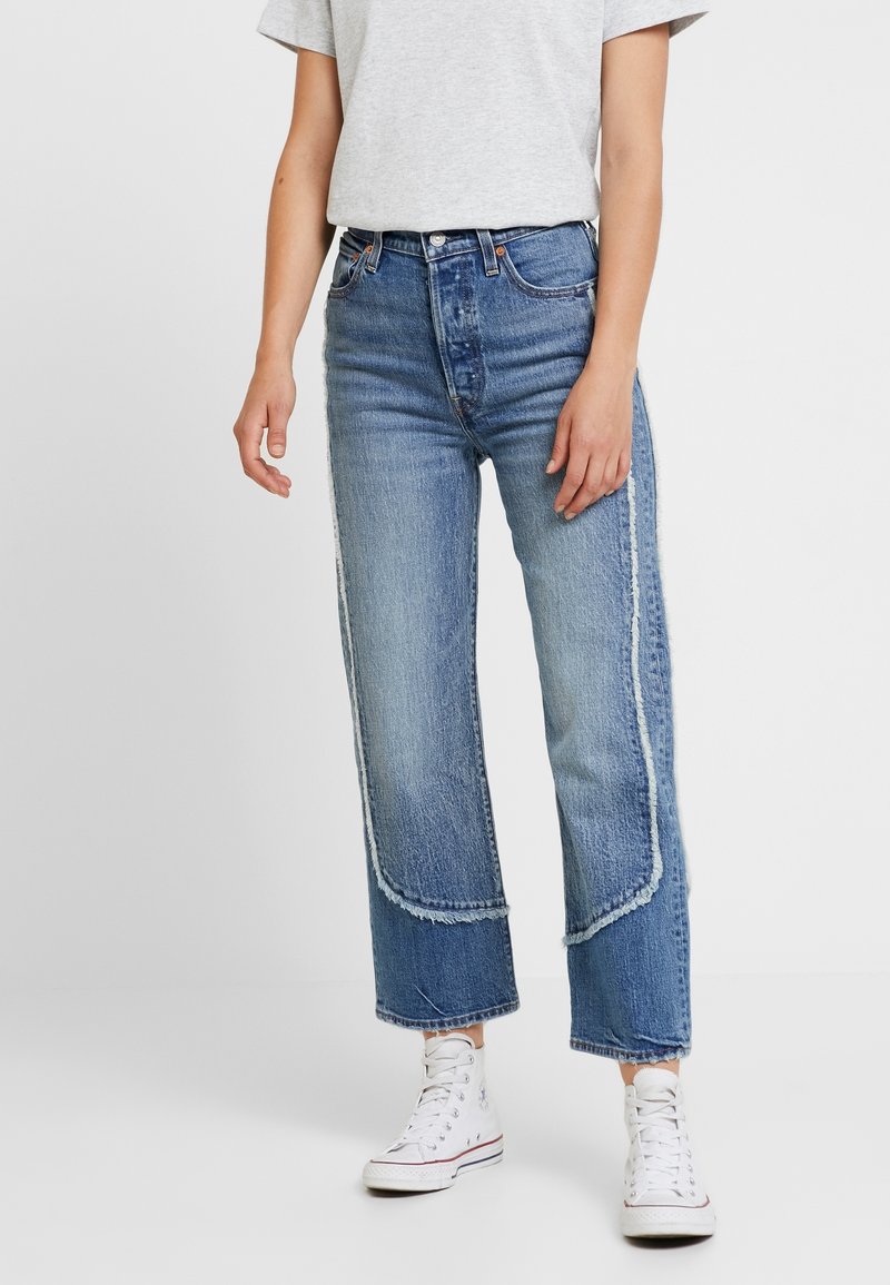 Jean en denim bleu clair taille haute avec bords effilochés, coutures contrastées et coupe décontractée, associé à des baskets en toile blanches.