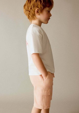 Young red-haired boy stands sideways with hands in pockets, wearing a white t-shirt and light pink shorts against a neutral background.
