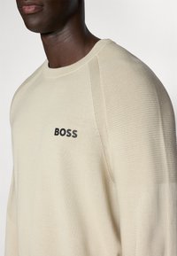 Beige Strickpullover mit gerippter Textur, mit Rundhalsausschnitt und dunklem "BOSS"-Logo auf der Brust. Nahaufnahme der Schulter.