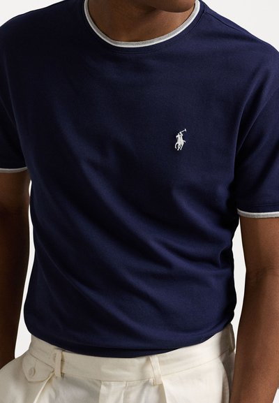 T-shirt de manga curta azul-marinho com decote arredondado, apresentando um acabamento cinza claro e um logótipo bordado branco no peito. Textura suave.