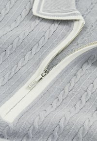 Pulover gri deschis cu model din tricot în cablu, având un închizător cu fermoar alb și accesorii de fermoar din argint. Textură moale și design clasic al gulerului.