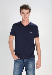 Navy blue cotton V-neck T-shirt featuring short sleeves and a small green logo on the left chest, paired with light blue jeans.