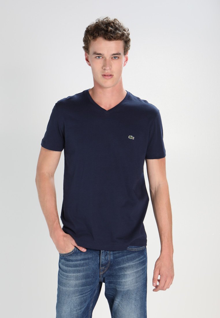 Navy blue cotton V-neck T-shirt featuring short sleeves and a small green logo on the left chest, paired with light blue jeans.
