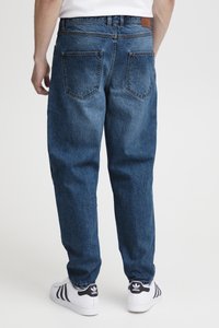 Egy személy laza szabású kék farmernadrágot és fehér Adidas tornacipőt visel, háttal áll a kamera felé egy egyszerű fehér háttér előtt.