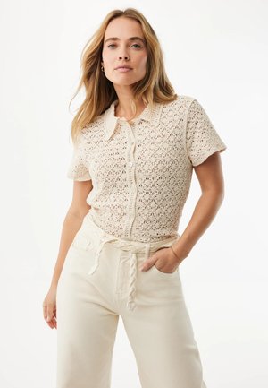 Kvinde iført en beige hæklede korte ærmer knap-op top og cremefarvede bukser med et flettet stofbælte, poserer foran en ens hvid baggrund.