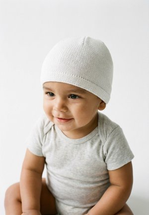 Lächendes Kleinkind, das eine weiße Strickmütze und einen hellgrauen kurzärmeligen Strampler trägt, sitzt vor einem schlichten weißen Hintergrund.