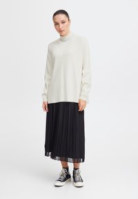 Pulover alb cu maneci lungi, cu dungi, fustă maxi plisată neagră și pantofi sport negri cu detalii albe. Design simplu și curat, cu culori contrastante.