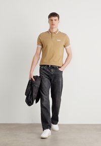 Hellbeige Polohemd mit weißen Streifen am Kragen, kombiniert mit dunklen Jeans und weißen Sneakers. Hält eine dunkle Jacke und steht vor einem neutralen Hintergrund.