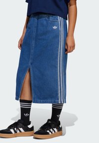Denim midi skjørt med en frontslisse, lyseblå farge, med hvite tre-stripes accenter og Adidas-logo. Kombinert med svarte joggesko og sokker.