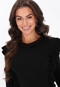 Czarny sweter ze wzorem prążkowym, z falbaniastym dekoltem i rękawami. Średniej grubości dzianina o gładkiej fakturze, dopasowany krój i długie rękawy.