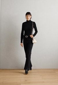 Schwarzes Rollkragenoberteil kombiniert mit schwarzen Schlagjeans, akzentuiert durch eine silberne Gürtelschnalle und einer kleinen weißen Umhängetasche.