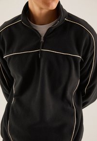 Mand iført sort fleece-pullover med hvid piping og halvdels lynlås krave, står med hænderne bag ryggen foran en ensartet baggrund.