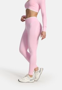Femme portant un crop top à manches longues rose et un legging taille haute assorti avec des baskets blanches, debout de profil devant un fond uni.