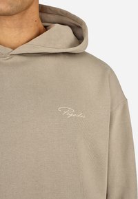 Beżowa bluza z kapturem o miękkiej fakturze, z przednią kieszenią i białym haftem z logo "Pegador" na piersiach. Regulowany kaptur na sznurki.