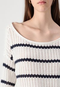 Suéter de punto blanco con franjas horizontales en navy, con un escote amplio y una textura suelta y aireada. Los detalles incluyen un diseño de tejido abierto.