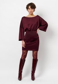 Bordeaux satijnen jurk met brede mouwen en een getailleerde taille, gecombineerd met doorschijnende zwarte bloemenvisnetpanty's en glanzende bordeaux kniehoge laarzen.