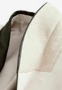 Col haut de blazer en feutre crème avec bordures sombres, doté d'une doublure intérieure lisse et de détails de surpiqûre contrastés ; design moderne et structuré.