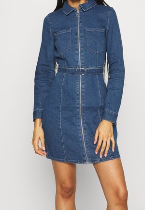 Robe en denim à manches longues, fermeture éclair à l'avant, ceinture à la taille et deux poches poitrine portée par une personne debout aux cheveux tressés.