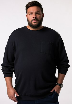 Dunkelblaues langärmliges Crewneck-Sweatshirt aus strukturiertem Stoff, mit einer Vordertasche und lockerer Passform, kombiniert mit blauen Jeans und einer Uhr.