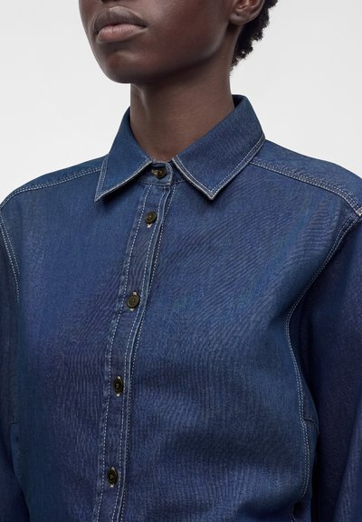 Personne portant une chemise en jean bleu foncé boutonnée, avec des surpiqûres blanches contrastantes et un col pointu, visible des épaules au menton.