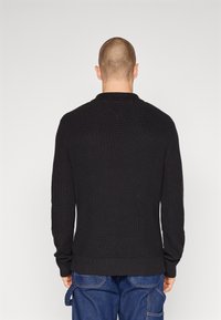 Černý pletený svetr s žebrovanou strukturou, kulatým výstřihem a dlouhými rukávy. Má přiléhavý lem a hladký zadní díl, nošený s modrými džínami.