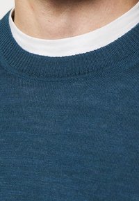 Blue knit sweater with a ribbed crew neckline, layered over a white shirt. Smooth texture with a subtle sheen.