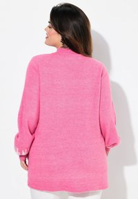 Flauschiger rosa Pullover mit hohem Kragen, überschnittenen Schultern und langen Ärmeln, die weiße Akzente an den Bündchen aufweisen. Glatte Textur und lockere Passform.