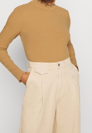 Kobieta ubrana w dopasowaną prążkowaną beżową bluzkę z długim rękawem, zdobioną falbankami przy szyi i mankietach, zestawioną z beżowymi szerokimi spodniami z wysokim stanem i kieszeniami.