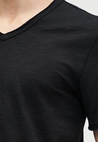 Κοντινό πλάνο μαύρου T-shirt με V λαιμόκοψη που φοριέται στον άνω κορμό ενός ατόμου, δείχνοντας την υφή του υφάσματος και τις λεπτομέρειες των ραφών.